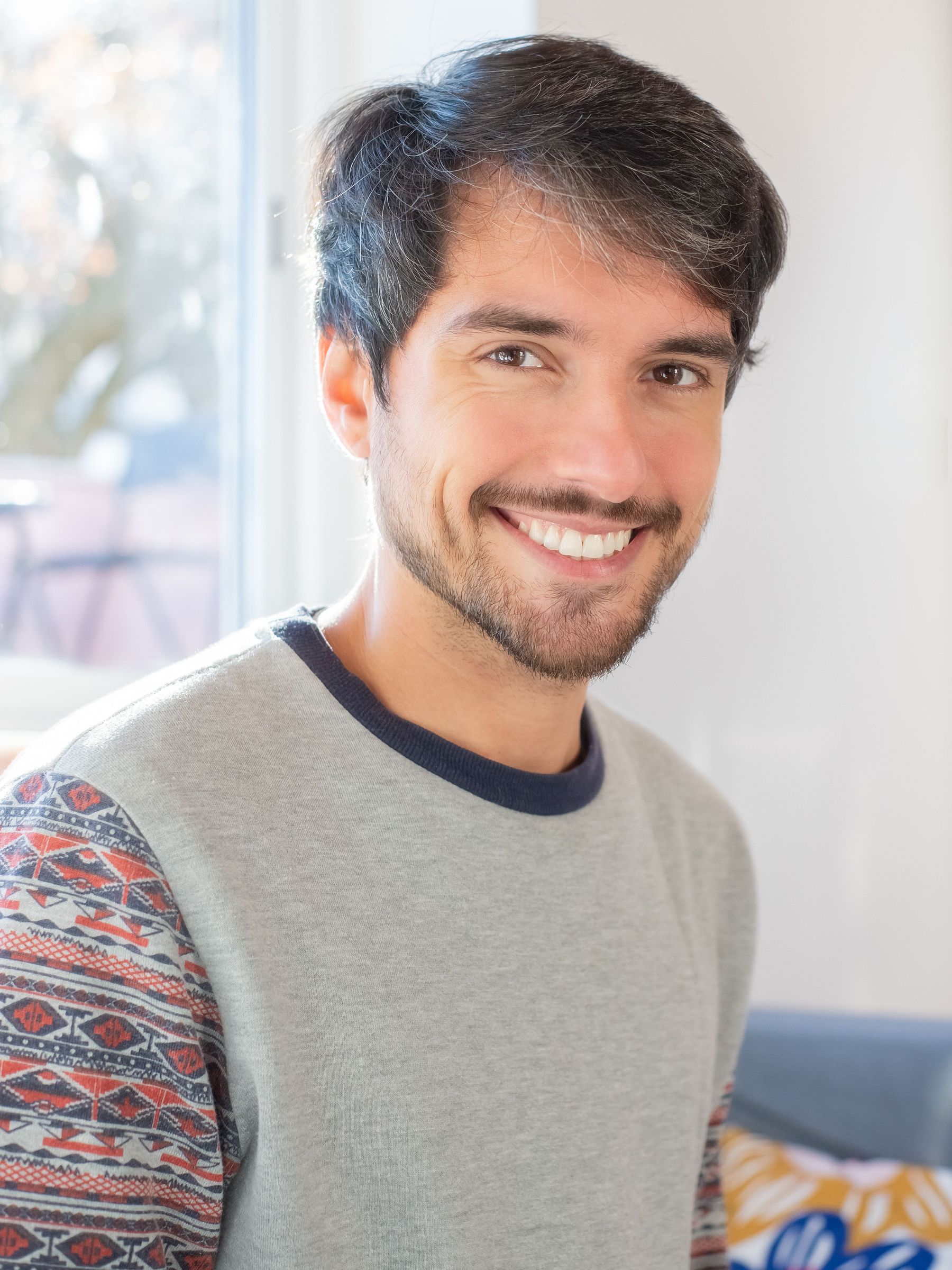Portret al lui Alex Marin, antrenor personal: bărbat cu barbă, zâmbet larg și autentic, privire directă către cameră, pulover casual gri cu mânecă cu model